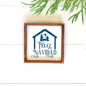 May include: A wooden sign with a white background and a blue silhouette of the nativity scene. The sign reads "Feliz Navidad" in blue lettering.
