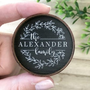 May include: A circular wooden ornament with a dark stained finish and a natural wood rim. The center features a white printed design of a leafy wreath surrounding the text "the ALEXANDER family" in a mix of script and serif fonts.