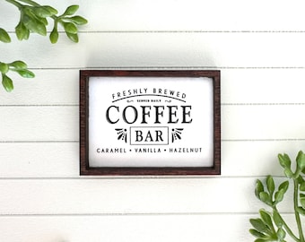 May include: A white wooden sign with a brown frame reads "Freshly Brewed Served Daily Coffee Bar Caramel Vanilla Hazelnut".