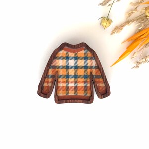 May include: A wooden brooch shaped like a sweater with a plaid pattern in shades of orange, blue, and white.