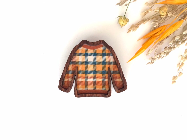 May include: A wooden brooch shaped like a sweater with a plaid pattern in shades of orange, blue, and white.