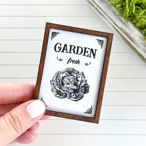May include: A brown wooden framed sign with white background and black text that reads "GARDEN fresh" with a black and white illustration of a head of lettuce.