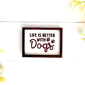 May include: A white wooden sign with a brown frame, featuring the text "Life is better with Dogs" in brown lettering with paw prints.
