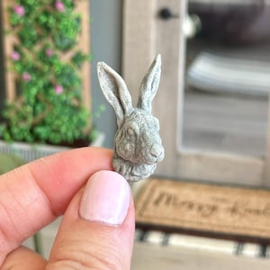 May include: A small, gray, ceramic rabbit head sculpture. The sculpture is detailed with realistic features, including the rabbit's ears, nose, and whiskers.