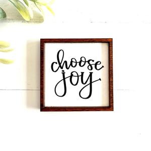 May include: A square wooden sign with a dark brown frame and a white center panel. The text "choose joy" is printed in black, cursive lettering in the middle of the sign. The sign features a clean, simple design with high contrast between the dark frame and the white background.