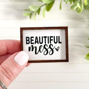 May include: A small, rectangular wooden sign with the words "BEAUTIFUL mess" in black script. The sign has a white background and a small heart symbol. The frame is a dark brown colour. Decorative home decor.