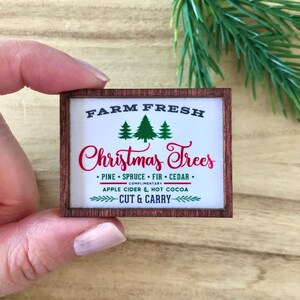 May include: A miniature wooden sign with the text "FARM FRESH Christmas Trees" in black and red. The sign includes green tree illustrations and mentions pine, spruce, fir, and cedar. It also mentions apple cider and hot cocoa.