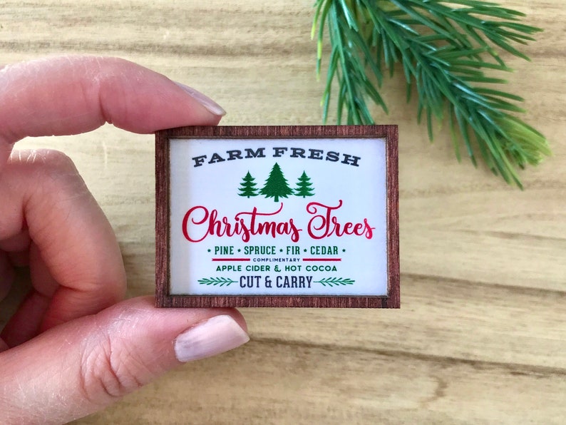May include: A miniature wooden sign with the text "FARM FRESH Christmas Trees" in black and red. The sign includes green tree illustrations and mentions pine, spruce, fir, and cedar. It also mentions apple cider and hot cocoa.