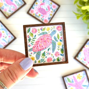 May include: A small square wooden frame containing a colorful illustration of a sea turtle surrounded by pink and yellow flowers and green foliage. The turtle has a pink patterned shell and blue speckled skin, set against a white background.