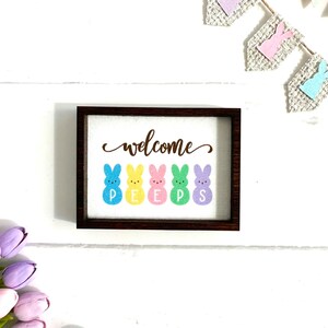 May include: A wooden sign with a brown frame and white background. The sign says "welcome" in brown cursive lettering and features five colorful Peeps marshmallow bunnies spelling out "PEEPS".