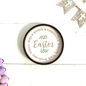 May include: A white round wooden sign with a brown wooden frame. The sign has the words "Easter", "three days", "miracle", "resurrection", "empty tomb", "redeemer" written in brown text around a green wreath.