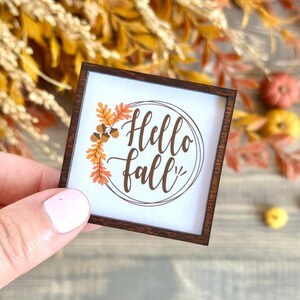 May include: A small, square wooden sign with a white background and the words "Hello Fall" in brown script. The sign is framed in dark brown wood and decorated with orange and brown fall leaves and acorns. The sign is held by a person.