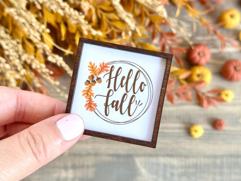 May include: A small, square wooden sign with a white background and the words "Hello Fall" in brown script. The sign is framed in dark brown wood and decorated with orange and brown fall leaves and acorns. The sign is held by a person.