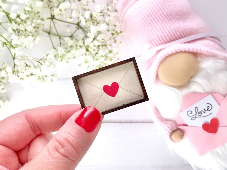 May include: A small wooden frame with a white envelope and a red heart inside. The frame is on a white surface. The word 'Love' is written on a pink envelope in the background.