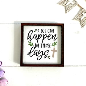 May include: A wooden sign with a white background and brown frame. The sign says "A lot can happen in three days." with a small cross next to the word "days".