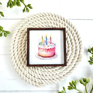 May include: A watercolour painting of a pink birthday cake with colourful sprinkles and candles in a brown wooden frame. The frame is on a white rope coaster.