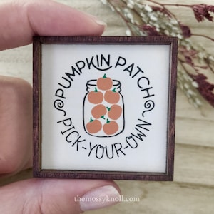 May include: A white wooden sign with a brown frame, featuring a circle with the text "Pumpkin Patch Pick-Your-Own" and a drawing of a mason jar filled with orange pumpkins.