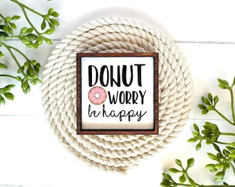 May include: A square wooden sign with a dark brown frame and a white background. The text reads DONUT WORRY be happy in black lettering, with a small illustration of a pink frosted donut with sprinkles replacing the letter O in donut.