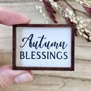 May include: A small wooden sign with a white background and a brown frame. The sign says "Autumn Blessings" in black cursive lettering.