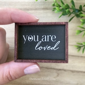 May include: A small wooden sign with a black background and white lettering that reads "you are loved" with a small white heart above the word "you". The sign has a dark brown frame.