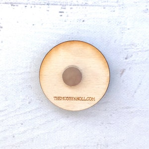 May include: A round wooden coaster with a small metal disc in the center. The coaster has the text "THEMOSSYKNOLL.COM" printed on the bottom.