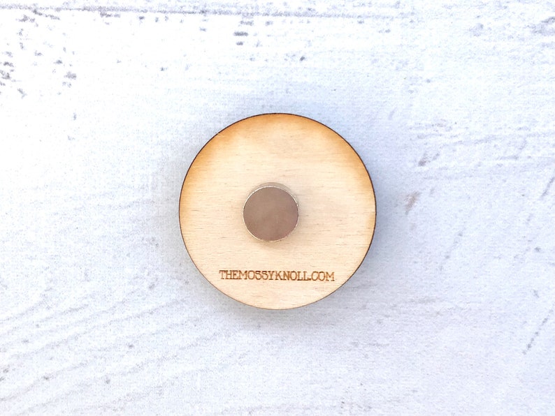 May include: A round wooden coaster with a small metal disc in the center. The coaster has the text "THEMOSSYKNOLL.COM" printed on the bottom.