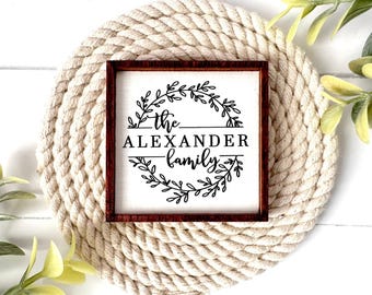 Puede incluir: Un letrero cuadrado de madera con un marco marrón oscuro y un centro blanco. El letrero presenta texto negro que dice "the ALEXANDER family" centrado dentro de un diseño de corona botánica circular hecho de hojas y ramas finas.
