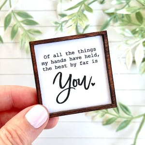May include: A small square wooden sign with a dark brown frame and a white center. The text printed in black reads: Of all the things my hands have held, the best by far is You, with a small heart symbol following the word You.