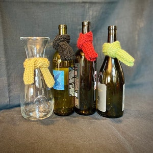 May include: Four glass bottles with knitted bottle cozies in black, red, yellow, and green. The bottles are arranged in a row on a gray surface.