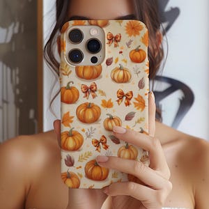 May include: A phone case featuring a repeating pattern of orange pumpkins, autumn leaves, and bows on a cream background. The case has a glossy finish and is held up in front of a person's face.
