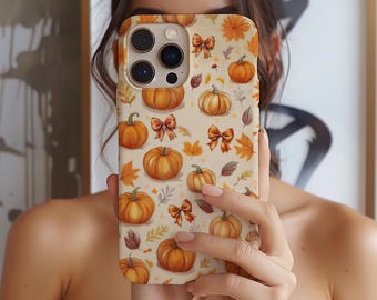May include: A phone case featuring a repeating pattern of orange pumpkins, autumn leaves, and bows on a cream background. The case has a glossy finish and is held up in front of a person's face.