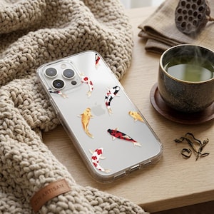 May include: Clear phone case featuring koi fish in red, black, and gold. The case is on a wooden surface next to a cup of tea and a knitted scarf. The case has a Zen Artisan label.