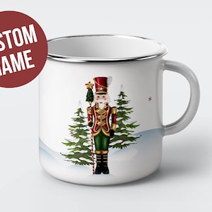 May include: White enamel mug with a silver rim and handle. The mug features a colorful illustration of a nutcracker standing in front of two pine trees with a snowy background.