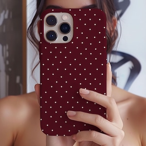 May include: A burgundy phone case with a white polka dot pattern. The case is held up in front of a person's face, showcasing the phone's camera lenses. The case is designed to fit a smartphone.
