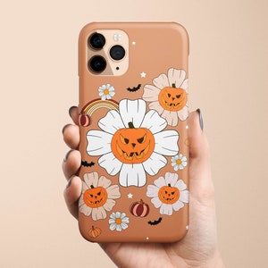 Pu&ograve; includere: Una custodia per telefono marrone a tema Halloween. La custodia presenta fiori bianchi con zucche arancioni all'interno. Le zucche hanno facce spettrali. Ci sono anche pipistrelli e stelle sulla custodia.