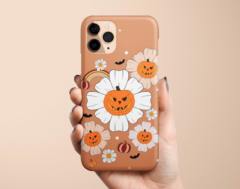 Pu&ograve; includere: Una custodia per telefono marrone a tema Halloween. La custodia presenta fiori bianchi con zucche arancioni all'interno. Le zucche hanno facce spettrali. Ci sono anche pipistrelli e stelle sulla custodia.