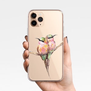 Puede incluir: Una funda transparente para teléfono con fondo dorado y una pintura de acuarela de dos pájaros rosados y verdes posados en una rama.