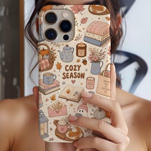 May include: A cream-colored phone case featuring a repeating pattern of autumn-themed illustrations including pumpkins, books, candles, tea, and leaves. The text COZY SEASON is printed in the center. The design includes soft pastel tones of pink, blue, and brown.