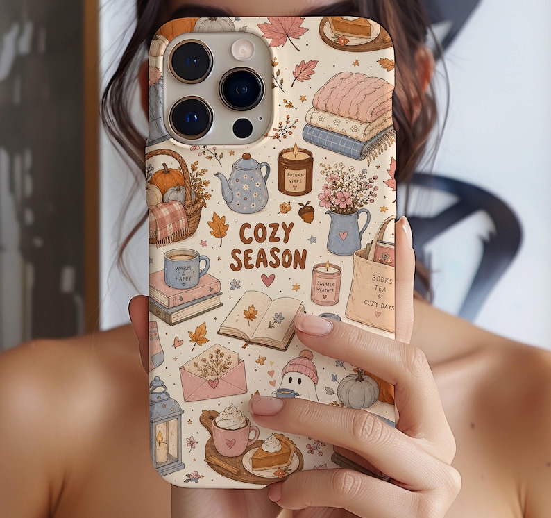 May include: A cream-colored phone case featuring a repeating pattern of autumn-themed illustrations including pumpkins, books, candles, tea, and leaves. The text COZY SEASON is printed in the center. The design includes soft pastel tones of pink, blue, and brown.
