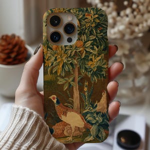 May include: A phone case with a tapestry design depicting a pheasant, trees, and foliage in brown, green, and yellow hues. The case has a camera cutout and is held in a person's hand.