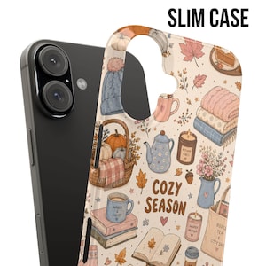 May include: A slim phone case featuring a cream background with a repeating pattern of autumn-themed illustrations including pumpkins, a knit hat, a basket, books, candles, and mugs. The text COZY SEASON is printed in brown. A black smartphone is visible behind.