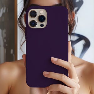 May include: A solid plum-colored phone case. The case has a smooth, matte finish and a camera cutout with multiple lenses. The phone case is held in front of a person's face.