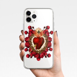 May include: A clear phone case with a design of a red heart with a crown of thorns and a cross, surrounded by red roses. The phone case is compatible with iPhone models.