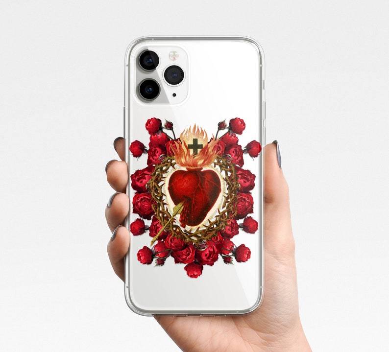 May include: A clear phone case with a design of a red heart with a crown of thorns and a cross, surrounded by red roses. The phone case is compatible with iPhone models.