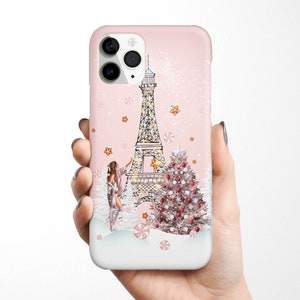 May include: A pink iPhone case with a white Eiffel Tower, a Christmas tree, and a woman decorating the tree. The background is a pink and white snowy scene.