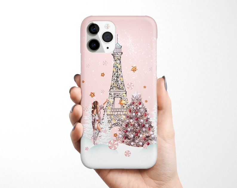 May include: A pink iPhone case with a white Eiffel Tower, a Christmas tree, and a woman decorating the tree. The background is a pink and white snowy scene.