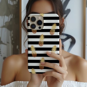 May include: A white phone case featuring black horizontal stripes and gold pineapple motifs. The case is held in front of a person's face. The phone case has a camera bump.