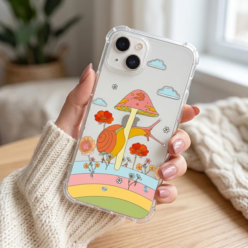 May include: Clear phone case with a colorful design featuring a snail, mushroom, flowers, and clouds. The design includes a rainbow-colored stripe at the bottom. The case is held in a person's hand.