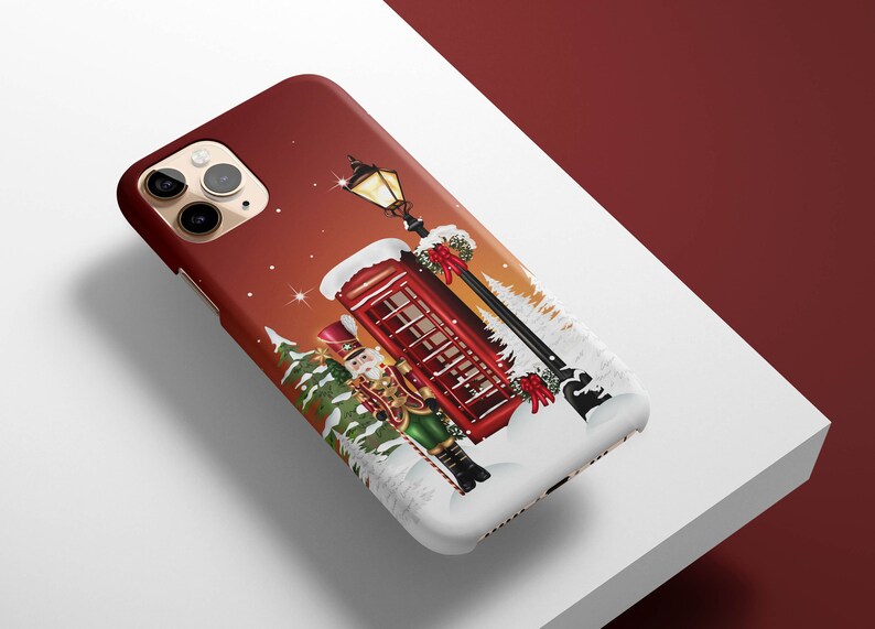 May include: A red phone booth with a snow-covered roof stands in a snowy winter scene. A nutcracker doll stands next to the phone booth, holding a candy cane. A street lamp with a lit lantern is behind the phone booth. The phone booth has a red door and windows. The scene is set against a red and orange background.