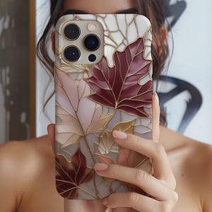 May include: A phone case featuring a stained-glass design with autumn leaves in shades of burgundy, gold, and cream. The case has a glossy finish and a protective bumper around the camera lenses.